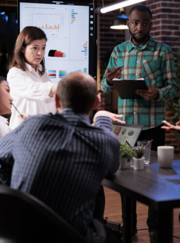 Caucasian business manager leading discussion in late night meeting talking about sales with team working overtime in startup office. Overworked business team brainstorming marketing strategy.